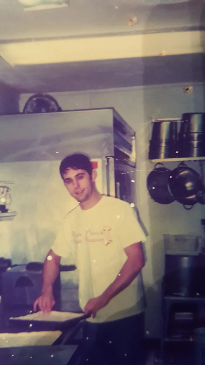 Archive photograph: a young man in a Picnic Pizza shirt working a Sicilian pan in the kitchen, pots hanging behind